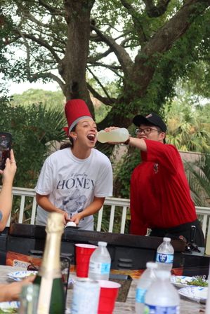 Outdoor backyard barbecue scene: a person in a red chef hat laughing as a cook in a red shirt pours lemonade into their mouth beside a griddle, picnic table with cups and water bottles in foreground and large trees in the background.