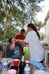 Friends laughing on a wooden porch surrounded by oak and palm trees at a summer gathering, one person playfully pours a drink from a bottle into another's mouth while a table holds red cups, water bottles and a champagne bottle.