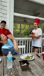 Playful summer scene on a screened backyard porch: woman wearing a tall red chef hat joyfully aims a colorful toy water gun at a seated woman who laughs and shields her face, while another person in a red chef coat smiles; wooden patio table with salad plate, water bottle and cups in foreground.