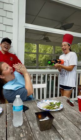 Playful summer scene on a screened backyard porch: woman wearing a tall red chef hat joyfully aims a colorful toy water gun at a seated woman who laughs and shields her face, while another person in a red chef coat smiles; wooden patio table with salad plate, water bottle and cups in foreground.