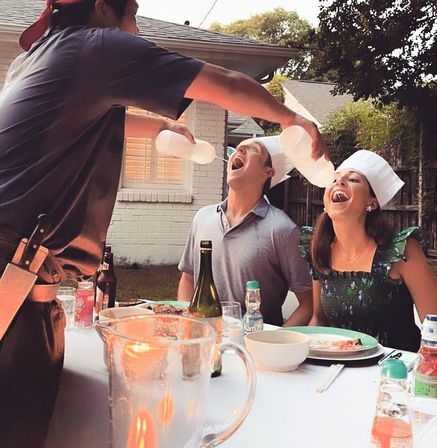 Playful backyard patio dinner with two guests in paper chef hats laughing as a server pours condiments from squeeze bottles into their open mouths, table set with plates, bottles and a pitcher.