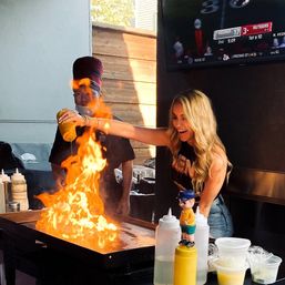 Woman laughing as she pours oil onto a flat-top grill, igniting a large flame at an outdoor teppanyaki-style station in a sports-bar setting