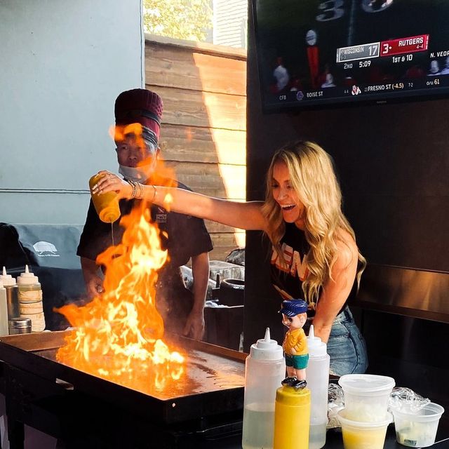 Smiling blonde woman pours oil onto a hot teppanyaki griddle, creating a dramatic blaze as a chef watches at an outdoor sports-bar style grill with condiment bottles and a game on TV in the background.