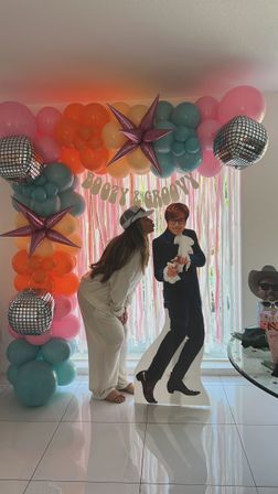 Fun indoor party backdrop with pastel pink, orange and teal balloon arch, metallic star and disco-ball balloons framing a pink fringe curtain and a "Boozy & Groovy" banner; a guest in white loungewear and a cap leans forward to kiss a life-size cardboard cutout of a retro-suited man on a glossy tiled floor.