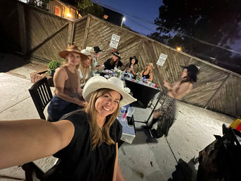 Smiling selfie of a woman in a white cowboy hat at a backyard evening cowboy-themed party, friends in hats gathered around a lit patio table by a wooden fence.