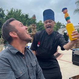 Outdoor patio: chef in black uniform and blue hat playfully sprays a thin stream of liquid from a novelty toy into a seated man's open mouth beside a garden fountain.