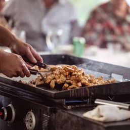 Sizzling bite-sized chicken being flipped with spatulas on a steaming flat-top griddle at a busy outdoor food station