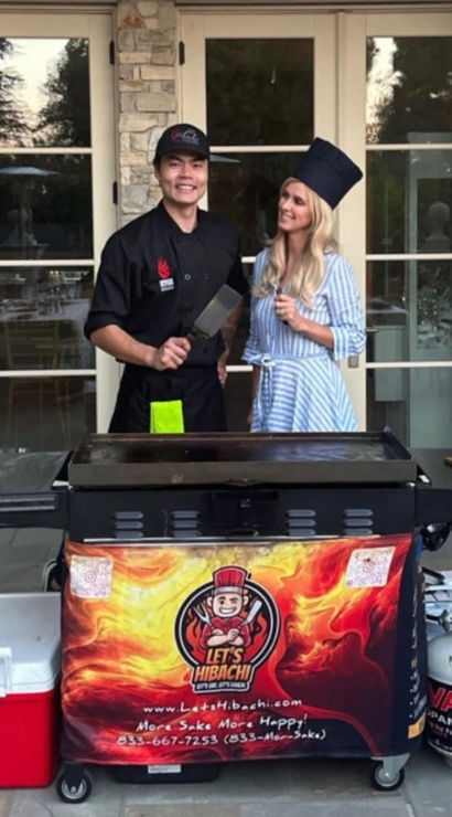 Outdoor hibachi scene on a patio: smiling chef in black uniform holding a spatula and a woman in a striped dress wearing a tall chef hat behind a flat-top griddle cart with condiment bottles, cooler, propane tank and colorful flame banner in front of glass doors and stone wall — backyard catering setup.