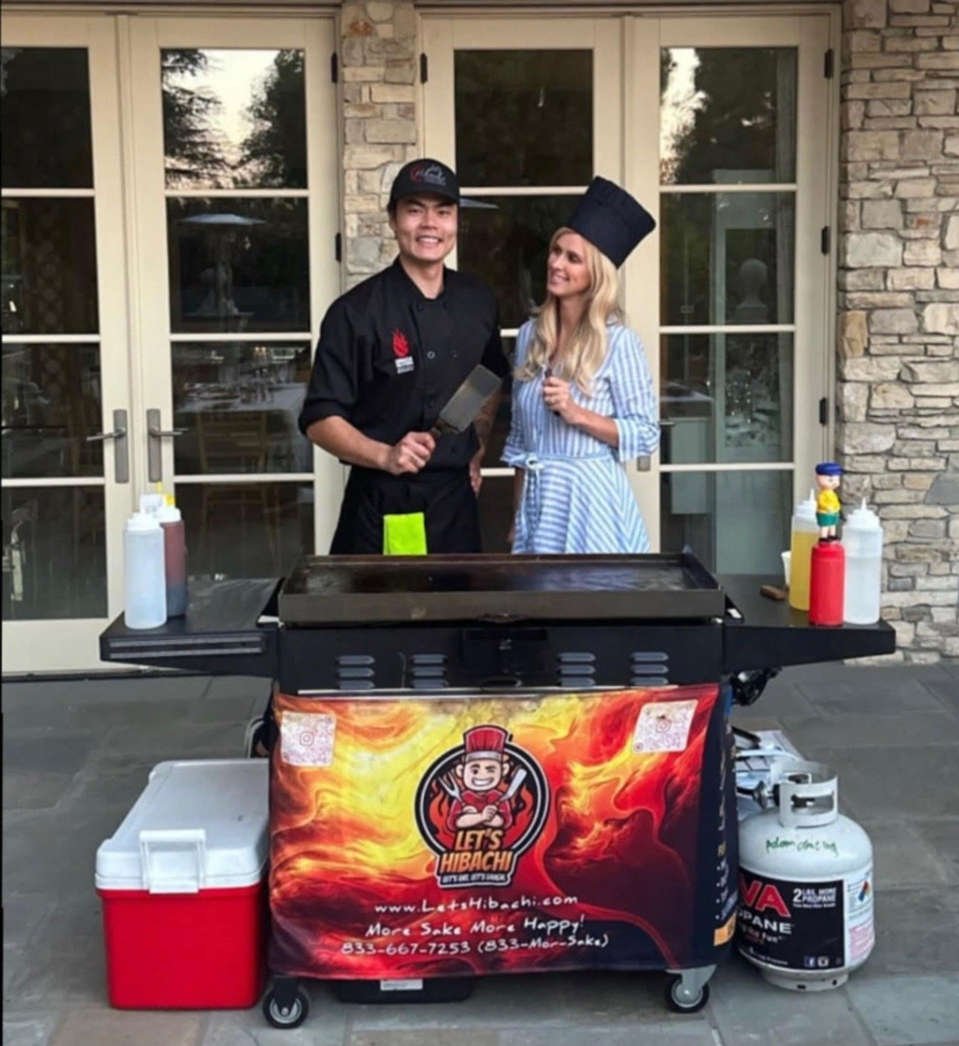 Outdoor hibachi scene on a patio: smiling chef in black uniform holding a spatula and a woman in a striped dress wearing a tall chef hat behind a flat-top griddle cart with condiment bottles, cooler, propane tank and colorful flame banner in front of glass doors and stone wall — backyard catering setup.