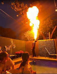 Person performing a dramatic fire‑breathing plume over a residential backyard patio pool at night, with string lights, potted plants, a wooden fence, and seated onlookers.