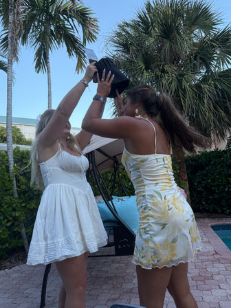 Two people in sundresses playfully holding a black bucket over one person’s head at a palm-tree lined poolside patio in a tropical backyard.
