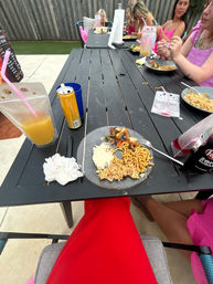 Backyard patio dining scene with a black slatted outdoor table holding plates of noodles, fried rice and grilled vegetables, drink pouches and cans, a paper towel roll, and a group of people in pink tops at the far end — casual summer meal.