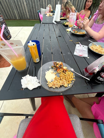 Backyard patio dining scene with a black slatted outdoor table holding plates of noodles, fried rice and grilled vegetables, drink pouches and cans, a paper towel roll, and a group of people in pink tops at the far end — casual summer meal.