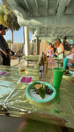 Tropical covered patio dining scene with palm trees, a long table draped in shiny gold foil, paper plates of mixed greens, green tumbler and champagne bottle, and guests serving food.