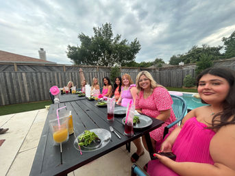 Backyard poolside outdoor dinner with a group of friends in pink outfits seated along a long black table, clear salad plates and colorful drinks, wooden fence, trees and cloudy sky above.