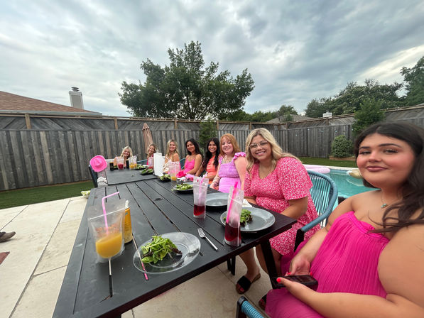 Backyard poolside outdoor dinner with a group of friends in pink outfits seated along a long black table, clear salad plates and colorful drinks, wooden fence, trees and cloudy sky above.