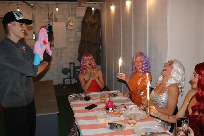 Friends at a backyard patio birthday party seated at a striped tablecloth, wearing colorful wigs and laughing as a host aims a pink water gun while others hold lit sparklers over dessert and drinks.