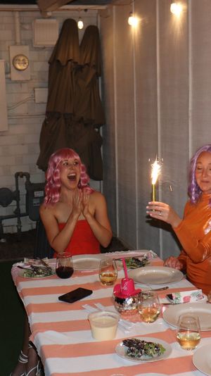 Friends at a backyard patio birthday party seated at a striped tablecloth, wearing colorful wigs and laughing as a host aims a pink water gun while others hold lit sparklers over dessert and drinks.