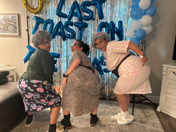Three friends in gray wigs and playful retro outfits twerking and posing in front of a metallic fringe backdrop and blue letter balloons at an indoor living-room party with a blue-and-white balloon column.