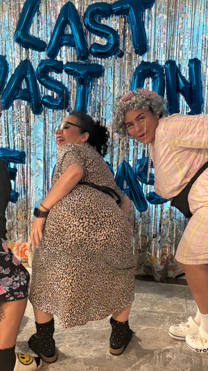 Three friends in gray wigs and playful retro outfits twerking and posing in front of a metallic fringe backdrop and blue letter balloons at an indoor living-room party with a blue-and-white balloon column.