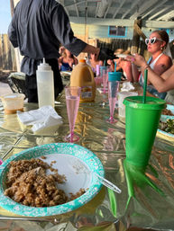 Outdoor patio brunch party with a gold foil-covered table, paper plate of rice, pink plastic champagne flutes, a green tumbler, orange juice jug and a server handing plates to seated guests.