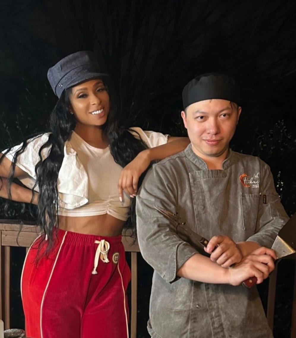 Smiling woman in a gray cap, white cropped top and red sweatpants poses beside a chef in a gray coat and black cap holding a spatula and tongs on a nighttime outdoor wooden deck.
