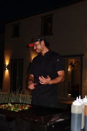 Chef in black coat and cap cooking mixed vegetables on a flat-top griddle at night on a modern residential patio, squeeze bottles and warm exterior lights in the background.