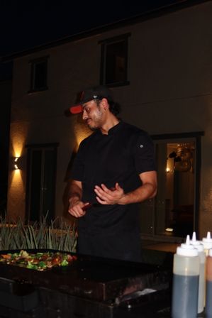 Chef in black coat and cap cooking mixed vegetables on a flat-top griddle at night on a modern residential patio, squeeze bottles and warm exterior lights in the background.