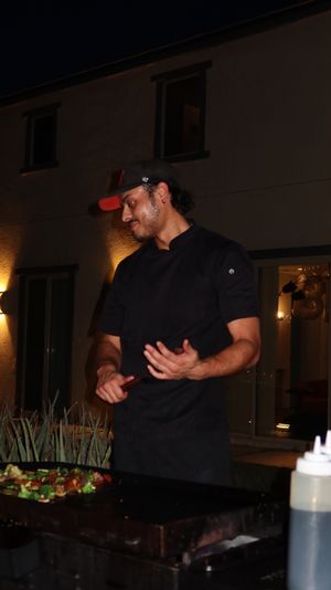 Chef in black coat and cap cooking mixed vegetables on a flat-top griddle at night on a modern residential patio, squeeze bottles and warm exterior lights in the background.