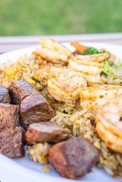 Close-up of grilled shrimp and seared steak cubes atop seasoned fried rice with corn, broccoli and carrots on a white plate with a soft outdoor green background