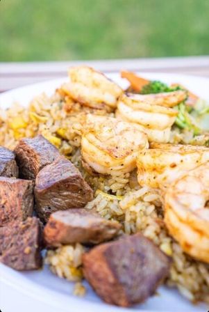 Close-up of grilled shrimp and seared steak cubes atop seasoned fried rice with corn, broccoli and carrots on a white plate with a soft outdoor green background
