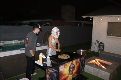 Backyard nighttime party scene: person in a silver costume and white wig cooking on a portable griddle while another guest watches, squeeze bottles of sauce on the table, hot tub in the background and a lit fire pit nearby under string lights.