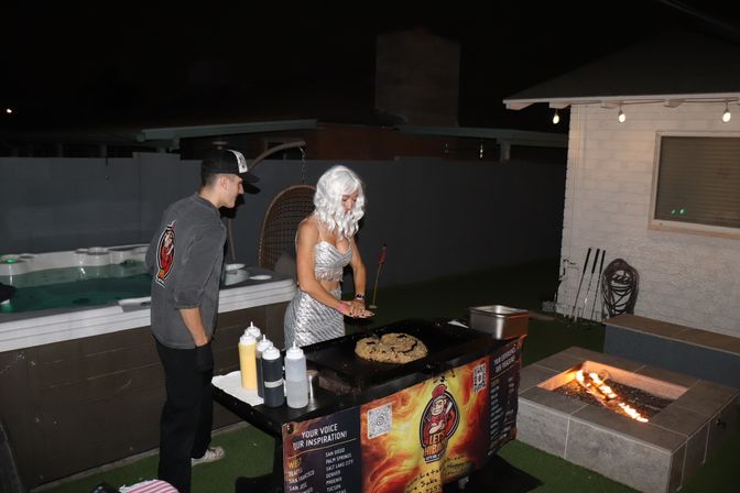 Backyard nighttime party scene: person in a silver costume and white wig cooking on a portable griddle while another guest watches, squeeze bottles of sauce on the table, hot tub in the background and a lit fire pit nearby under string lights.