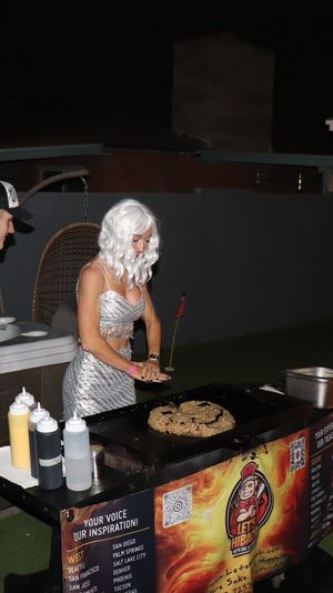 Backyard nighttime party scene: person in a silver costume and white wig cooking on a portable griddle while another guest watches, squeeze bottles of sauce on the table, hot tub in the background and a lit fire pit nearby under string lights.