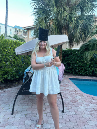 Person in a white sundress wearing a tall chef’s hat and holding a cleaver, posing poolside on a tropical patio with palm trees, lounge swing and tiled pavers.