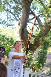 Excited woman on a sunlit porch holding a long flaming sword aloft during an outdoor backyard fire performance, with oak trees and lush greenery in the background.