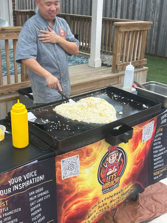 Cook preparing a large mound of fried rice on a hibachi-style griddle at an outdoor patio food booth, with squeeze bottles and a flame-themed banner