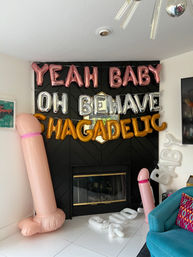Bachelorette party decorations in a living room: rose-gold "YEAH BABY" and silver "OH BEHAVE" foil balloons over a black fireplace, gold slogan balloons, two large inflatable pink novelty penises and scattered white letter balloons on white tile floor.
