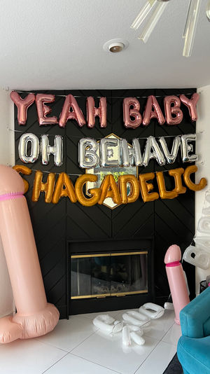 Bachelorette party decorations in a living room: rose-gold "YEAH BABY" and silver "OH BEHAVE" foil balloons over a black fireplace, gold slogan balloons, two large inflatable pink novelty penises and scattered white letter balloons on white tile floor.