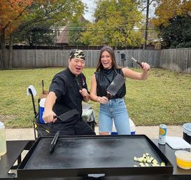 Two cheerful cooks posing with spatulas and a cleaver beside a large flat-top griddle at a suburban backyard cookout, diced zucchini on the hotplate and autumn trees in the background.