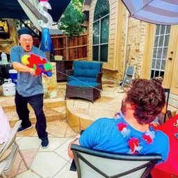 Summer backyard patio scene: person playfully firing a bright multicolor water gun at a seated friend wearing a red-white-blue lei, with wicker patio chairs, umbrella and brick house in the background.