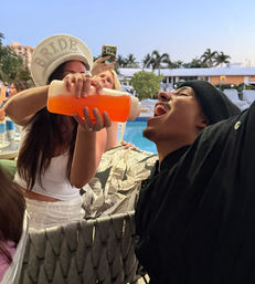 Poolside bachelorette scene: woman wearing a 'BRIDE' hat pours a bright orange drink from a squeeze bottle into a man's open mouth beside a resort pool with palm trees at dusk.