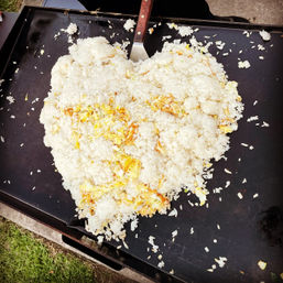 Overhead close-up of a heart-shaped pile of fried rice with scrambled egg on a black flat-top griddle, metal spatula at the top and grass visible at the edge — outdoor cookout.