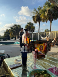 Poolside backyard party with palm trees and hammock, chef at a mobile grill serving a person wearing a veil, sunny afternoon, table with drinks and condiments.