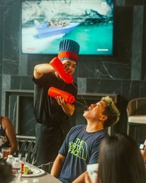 Restaurant server in a blue chef hat double-pours from red squeeze bottles into a diner’s open mouth during a lively table-side performance, with plates, drinks and a TV showing a boat in the background.