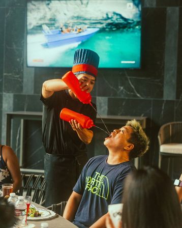 Restaurant server in a blue chef hat double-pours from red squeeze bottles into a diner’s open mouth during a lively table-side performance, with plates, drinks and a TV showing a boat in the background.