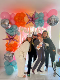 Three friends posing at an indoor party photo booth under a colorful balloon arch of pink, orange, teal and peach balloons with mirrored disco-ball and metallic star balloons, pink streamer backdrop, and a life-size cardboard cutout in a retro suit.