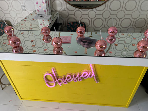 Bright yellow beauty bar counter with mirrored countertop, pink sequin double-bubble cups with straws, a handheld face fan, geometric wallpaper and neon pink cursive sign.