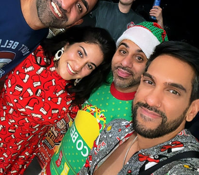 Nighttime house holiday party selfie of five friends in festive pajamas and ugly Christmas sweaters — one wearing a Santa hat, cars visible outside.