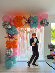 Indoor party photo backdrop with pastel balloon arch (pink, orange, yellow, teal), metallic purple star balloons and mirrored disco-ball balloons over pink fringe curtains and a "Boozy & Groovy" banner, with a retro-suited cardboard cutout prop on glossy white tile.